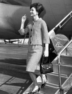 Jackie Kennedy en tailleur élégant pied-de-poule, descendant de l'avion à l'aéroport de New-York. Elle est coiffée d'un chignon chic et arbore un grand sourire. 