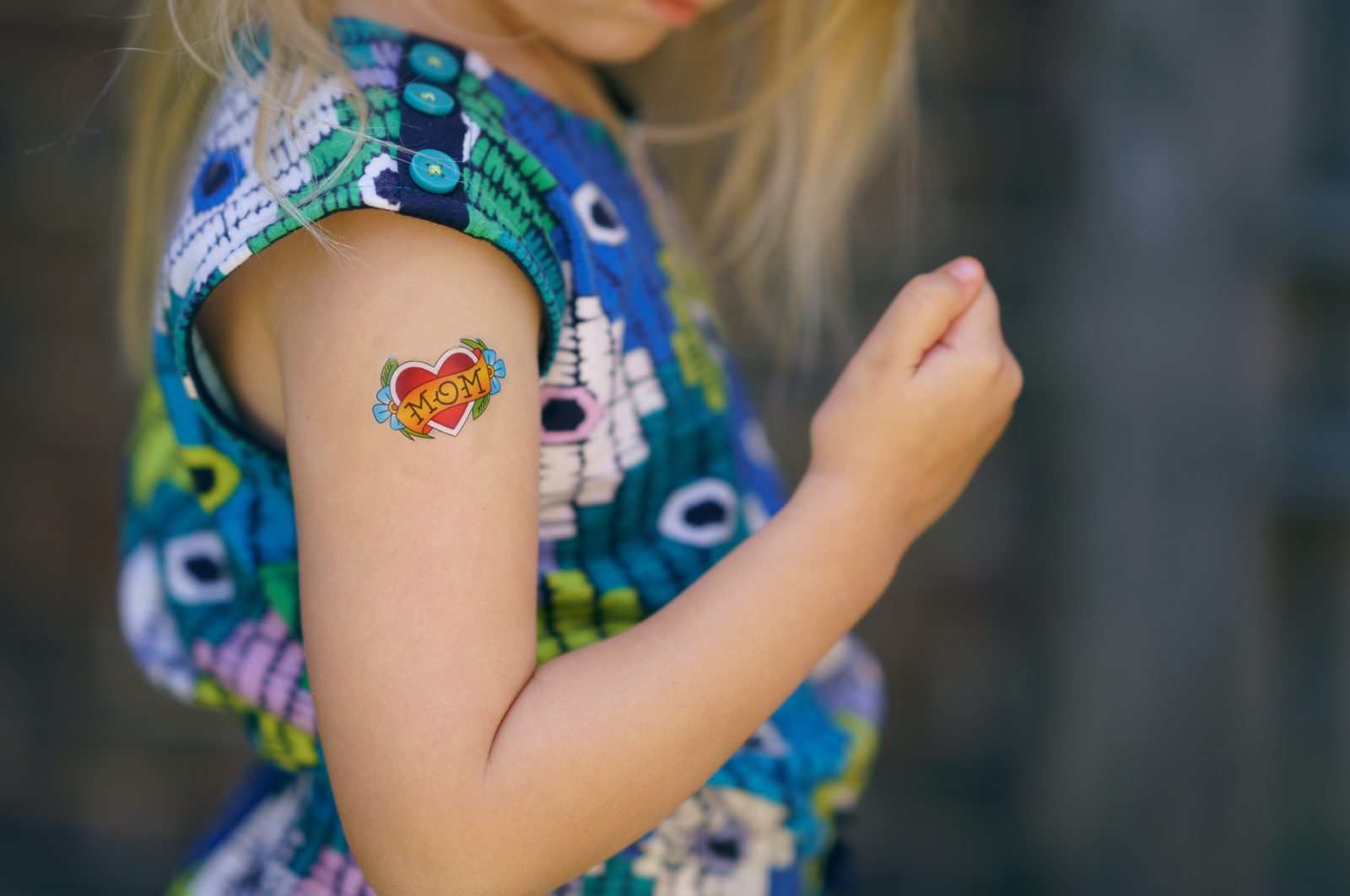 Une enfant qui porte un tatouage éphémère sur son bras droit. Celui-ci représente un cœur rouge avec des fleurs et un bandeau où est écrit "Mom".