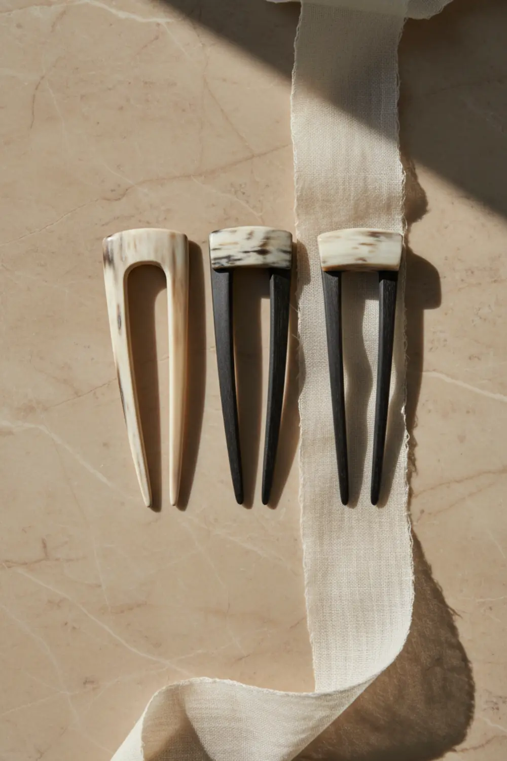 Épingles en U en corne polie et ruban de soie de mûrier drapés sur un bloc de marbre beige sous une douce lumière d'atelier.