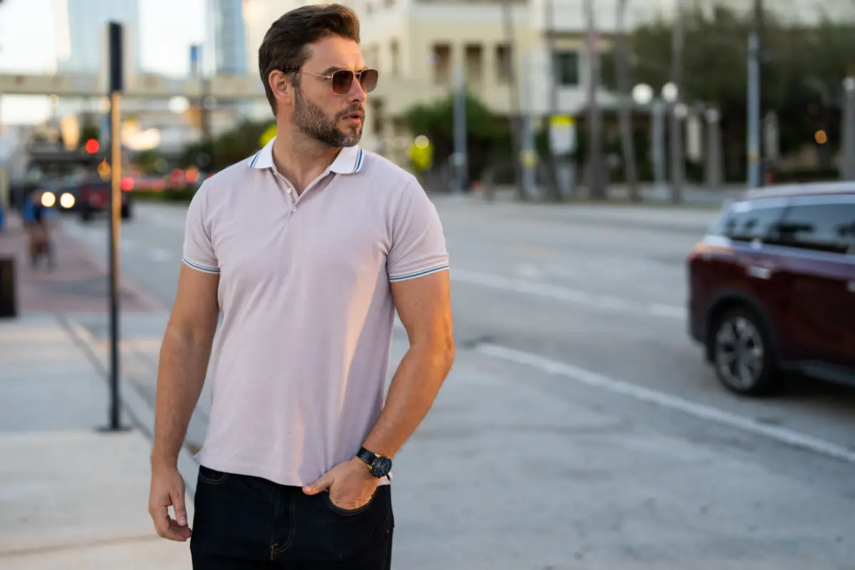 Homme portant un polo rose clair, des lunettes de soleil et un jean sombre en ville