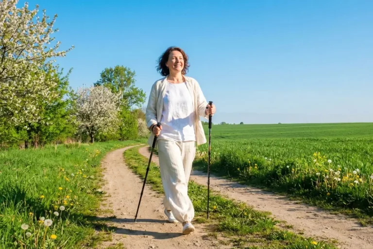 Femme de 50 ans pratiquant la marche nordique seule sur un chemin dégagé sous un ciel bleu vif au printemps, posture énergique et bâtons en mouvement.