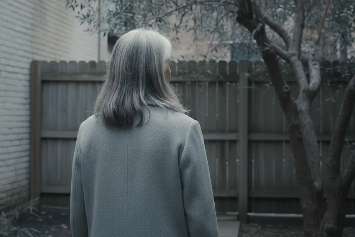 Femme de 50 ans vue de dos dans un jardin urbain, regardant un arbre, lumière froide et maîtrisée.