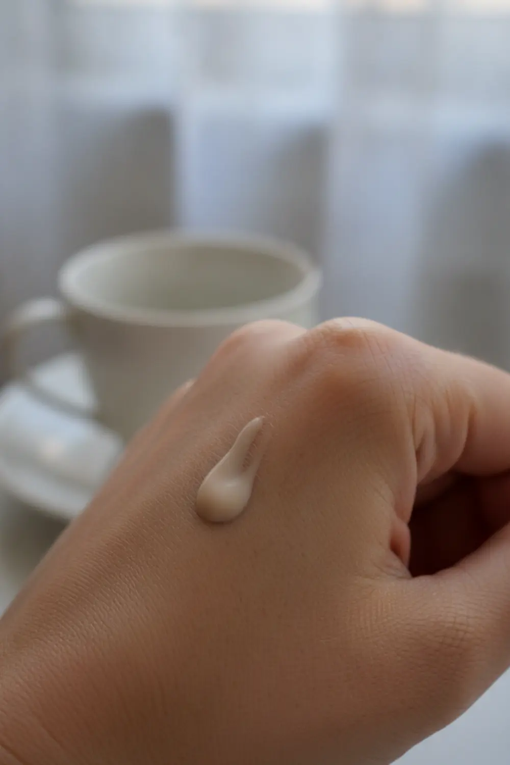 Goutte de BB crème déposée sur le dos d’une main, texture crémeuse et légère, fond flou avec tasse de thé et fenêtre.