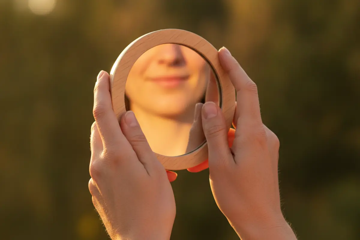 Mains féminines tenant un miroir rond avec un sourire en reflet, fond végétal flou, style beauté naturel