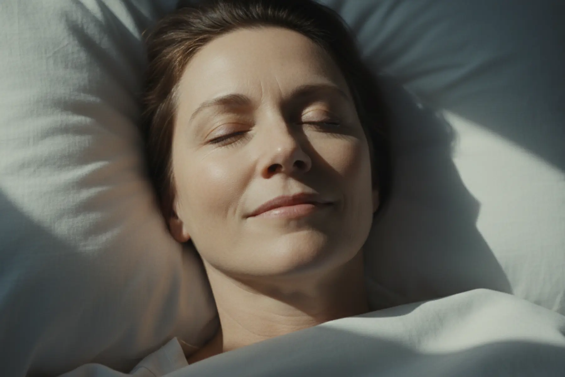 Rêves lucides : pourquoi s'offrir une nuit de conscience fait du bien au corps et à l'esprit 1 Femme d'une cinquantaine d'années allongée sur un drap de lin blanc, sourire apaisé, lumière du matin. Rêves lucides.