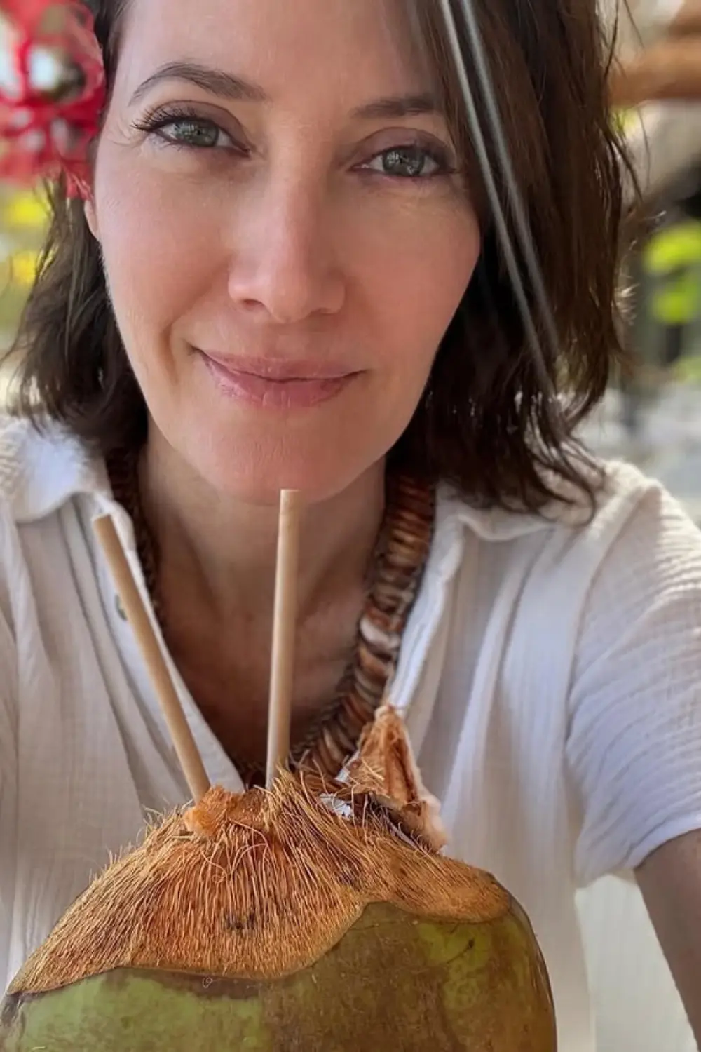 Gros plan Mareva Galanter souriante avec une fleur à l'oreille, tenant une noix de coco ouverte pour boire.