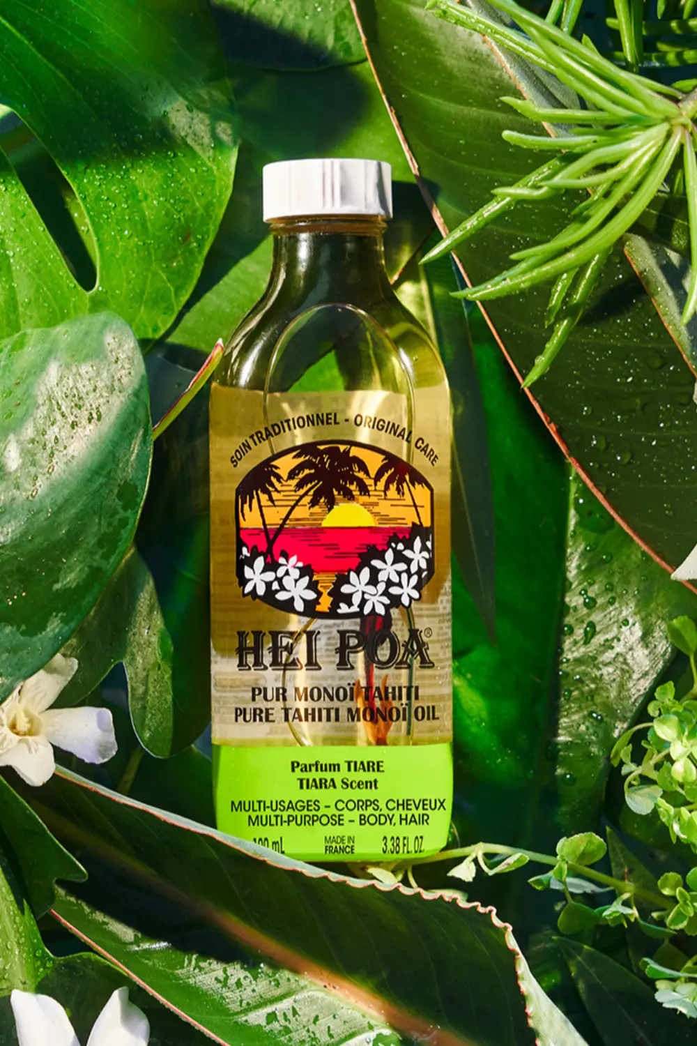 Flacon d'huile de monoï de Tahiti Hei Poa au parfum Tiaré, entouré de feuilles tropicales humides.