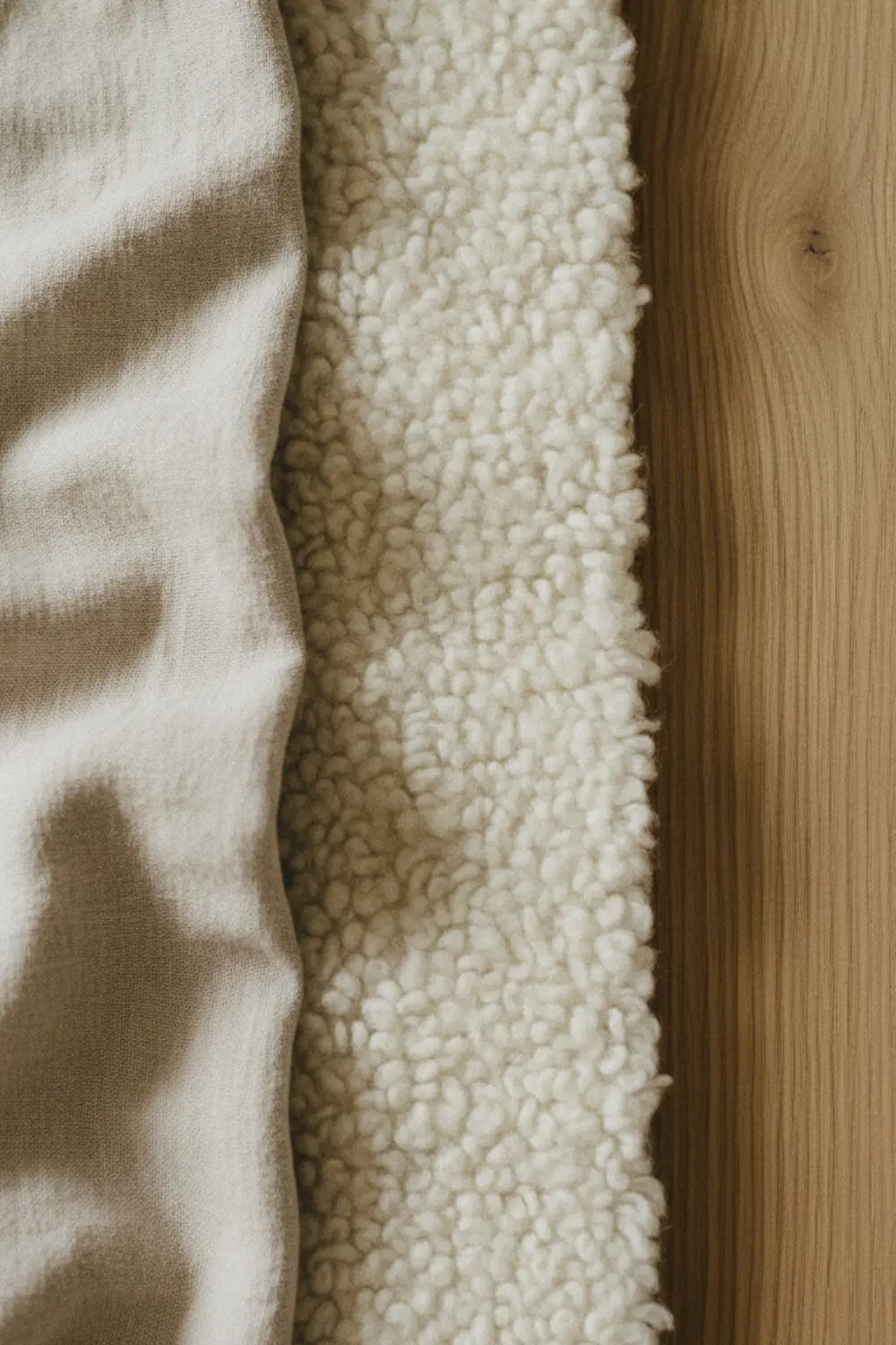 Détail tactile de trois matières naturelles : lin lavé froissé, laine bouclée ivoire et chêne brut à grain visible — textures warm minimalism