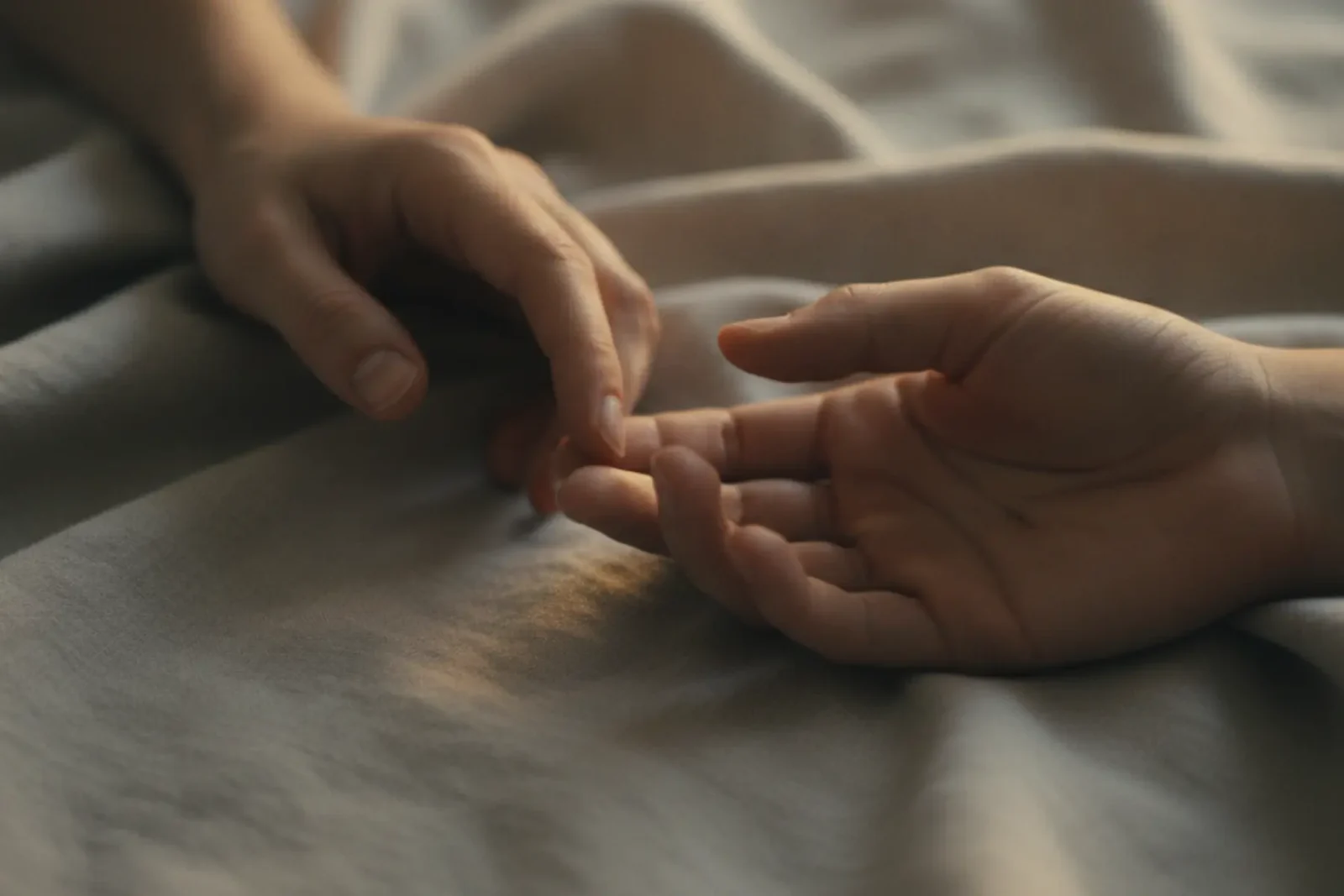 Gros plan artistique de deux mains qui se frôlent sur un drap en lin grège, lumière chaude, illustration du slow sex et du toucher sensoriel