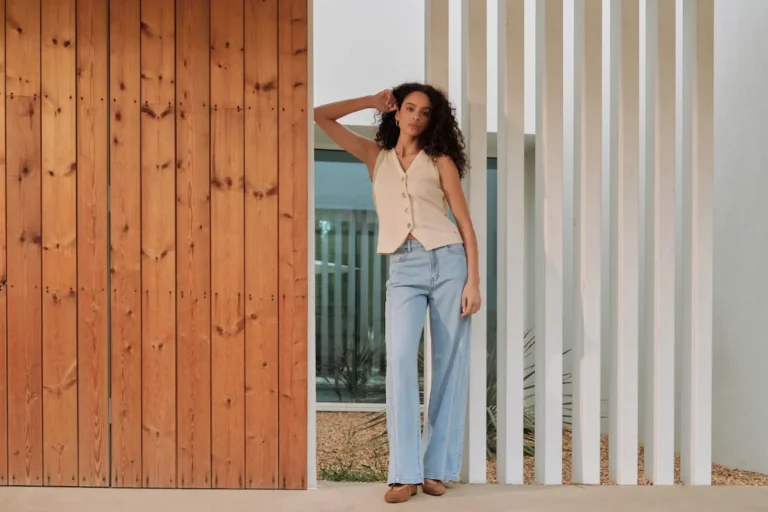 Comment adopter la tendance du denim fluide pour un look chic et décontracté ? 2 Femme portant un jean large en denim fluide clair et un gilet sans manches beige devant une architecture moderne en bois et colonnes blanches.