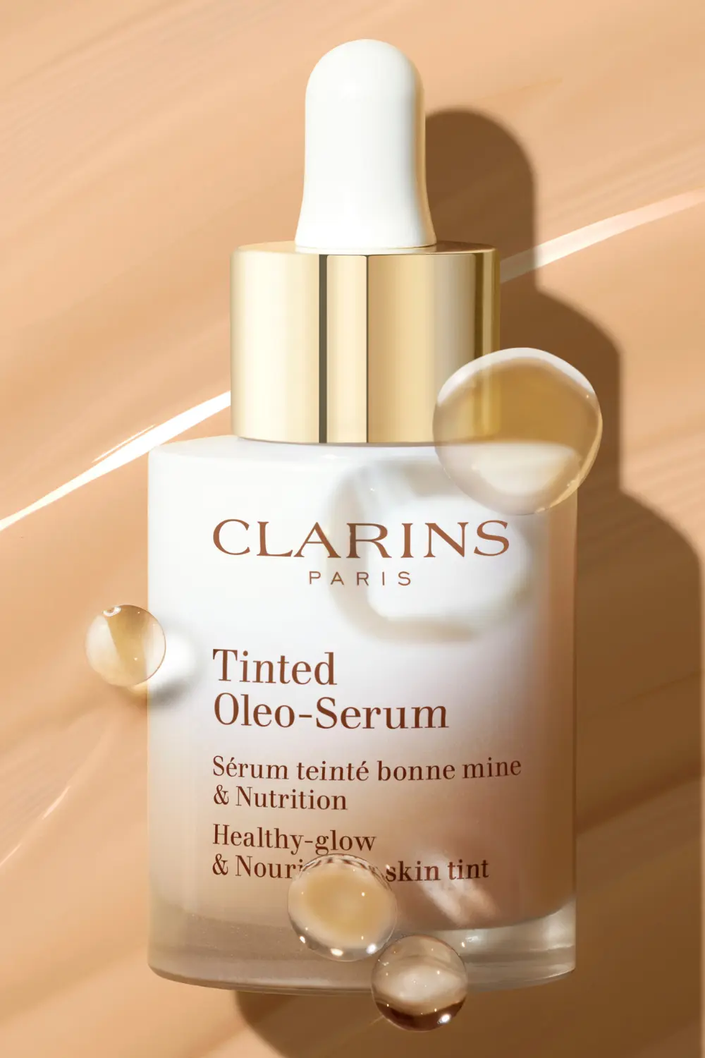Flacon de sérum teinté Tinted Oleo-Serum Clarins entouré de gouttes d'huile sur une texture de fond de teint.